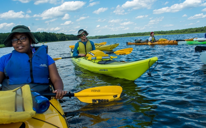 a group of teens take a break from paddling kayaks on a large body of water lined by trees